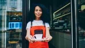 Woman coffee shop owner displaying a cup of coffee towards camera.